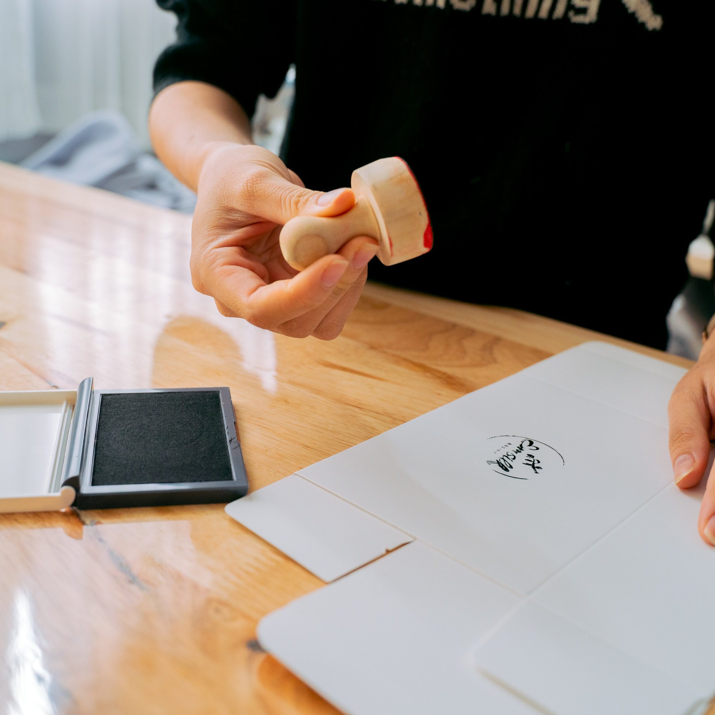 Hands using a stamp on paper at a wooden table, emphasizing craftsmanship.